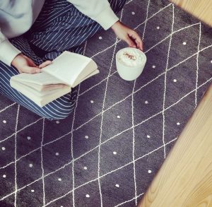 Donna seduta su un tappeto kilim orientale moderno grigio a rombi e puntini bianchi che legge un libro con una tazza di cappuccino